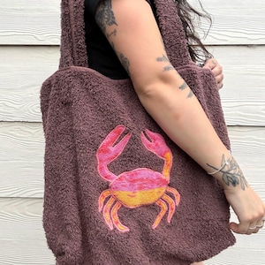 Puede incluir: Bolso tote de color malva oscuro con un diseño de cangrejo bordado. El cangrejo presenta colores rosa y naranja. El bolso tiene una textura suave y correas largas para llevar al hombro.