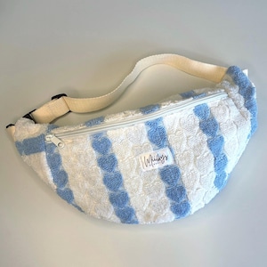 Pode incluir: Cinto de cintura listrado em branco e azul com uma alça ajustável creme e fecho de zíper. A bolsa tem uma aparência macia e texturizada e apresenta o texto "Whilkys" em um pequeno rótulo.