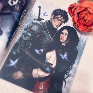 Può includere: Copertina di un libro fantasy con un'illustrazione di un uomo e una donna con capelli scuri e pelle chiara. L'uomo indossa un'armatura e una collana, e la donna indossa un mantello scuro. Sono raffigurate anche farfalle blu e una spada.