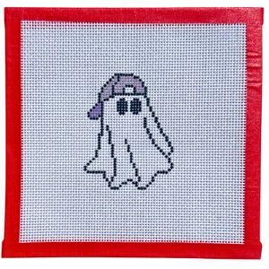 May include: A square needlepoint canvas featuring a white ghost character wearing a light purple baseball cap. The ghost is outlined in dark blue stitches with two dark blue circular eyes. The canvas is bordered by a bright red fabric tape.