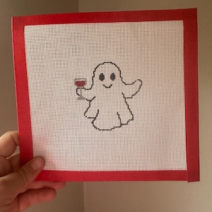 May include: A cross-stitch design featuring a smiling ghost holding a glass of red wine. The ghost is outlined in black on a white background. The design is framed with a red border.