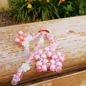 May include: A handmade beaded keychain featuring a pink flower and star design. The keychain includes pink, clear, and iridescent beads, with a red keyring. The flower and star are made with white and pink beads.