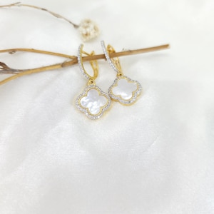 May include: Gold-coloured hoop earrings with a clover-shaped pendant. The pendant has a white, mother-of-pearl centre, framed by small, sparkling crystals. The earrings are presented on a white, satin-like surface, creating a luxurious feel.