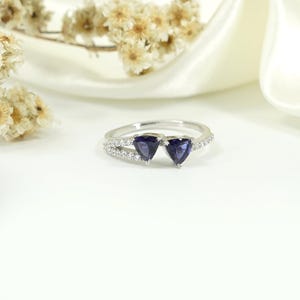 Może przedstawiać: Srebrny pierścionek z dwoma szafirowymi kamieniami w kształcie serca. Obręcz jest podzielona, z jednej strony ozdobiona małymi, przezroczystymi kamieniami. Pierścionek jest umieszczony na białym tle z delikatnymi kwiatowymi akcentami.