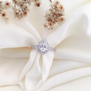 May include: A silver engagement ring with a pear-shaped center stone, surrounded by smaller accent stones. The ring is set on a white fabric, with dried flowers in the background. The ring's band is also adorned with small stones.