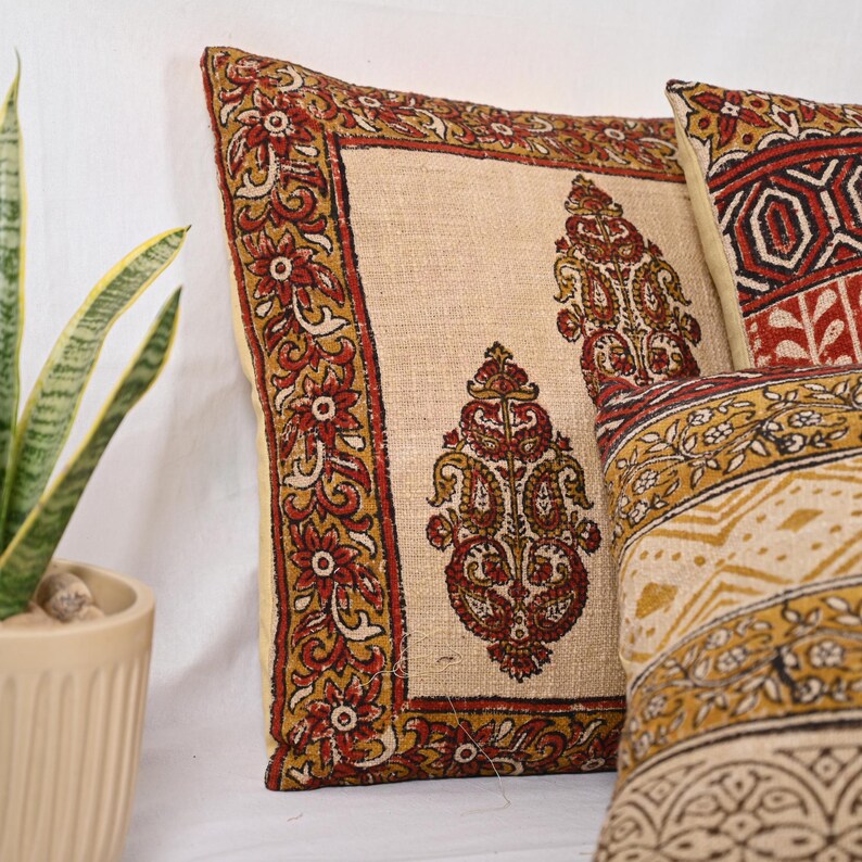 May include: Decorative throw pillows with a beige background and intricate patterns in red, gold, and brown. The pillows feature floral and paisley designs, adding a touch of elegance to any living space. The pillows are square-shaped.