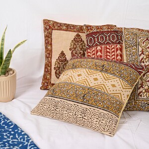 May include: Four decorative throw pillows with intricate patterns in shades of beige, gold, and burgundy. The pillows feature geometric and floral designs, creating a warm and inviting aesthetic. The pillows are arranged on a white surface.