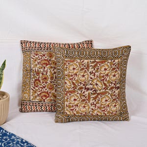 May include: Two square throw pillows with floral patterns. The pillows feature a cream-colored background with a repeating pattern of yellow and red flowers and leaves. The borders are decorated with geometric designs in black, yellow, and red.