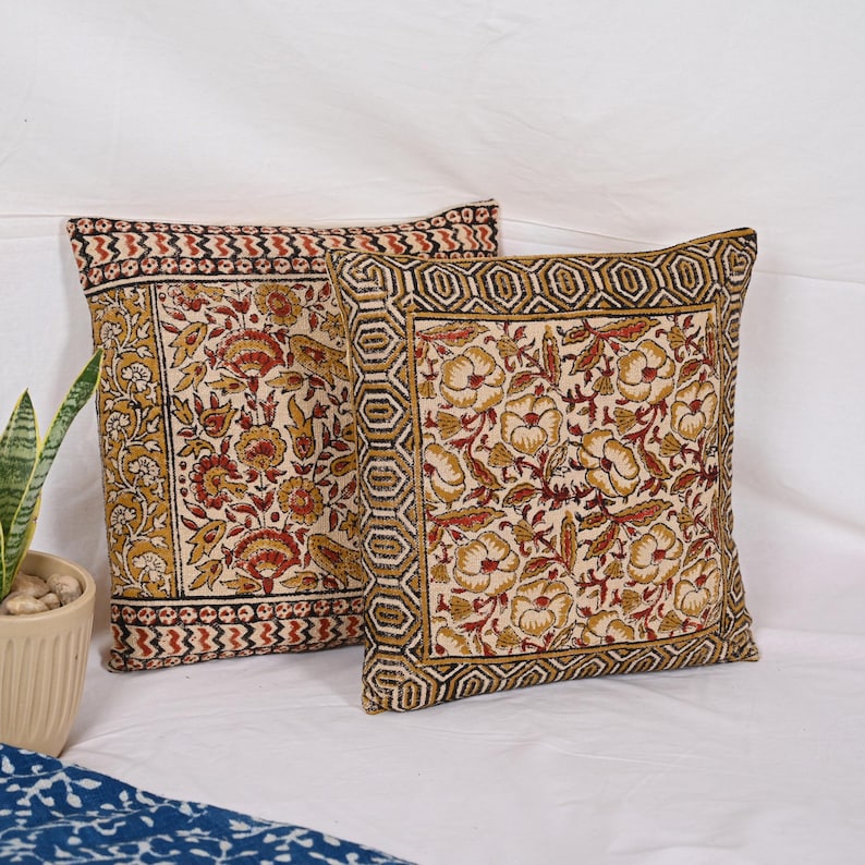May include: Two square throw pillows with floral patterns. The pillows feature a cream-colored background with red and gold floral designs. The borders have geometric and patterned designs in red, gold, and black.