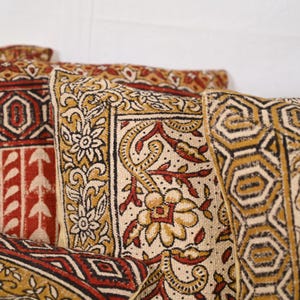 May include: Close-up of several decorative throw pillows with intricate patterns. The pillows feature a cream-colored background with floral and geometric designs in shades of red, gold, and black. The fabric appears to be textured.