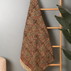 Könnte beinhalten: Gemustertes Textil mit sich wiederholendem floralem Design in Rot-, Grün- und Brauntönen. Der Stoff ist über eine Holzleiter drapiert und zeigt seine komplizierten Details und den Fransenrand. Das Textil ist wahrscheinlich ein dekorativer Artikel.