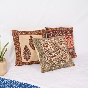 May include: Three decorative throw pillows with intricate floral and paisley patterns in shades of red, beige, and gold. The pillows are square-shaped and rest on a white surface, showcasing their detailed designs.