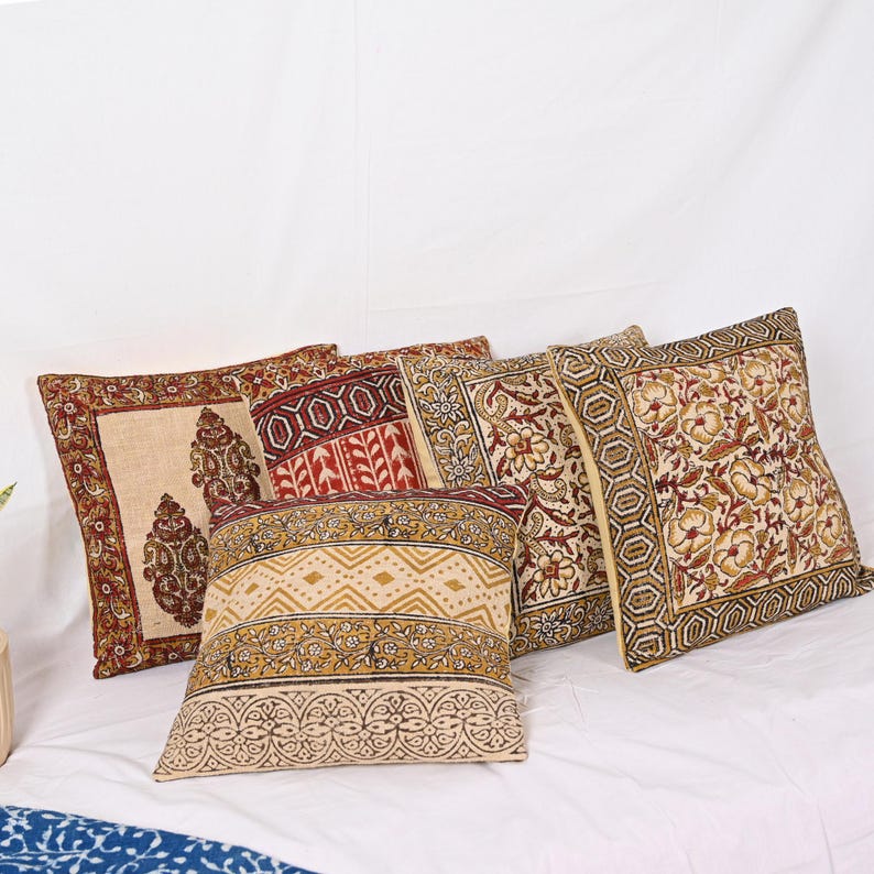May include: Five square throw pillows with intricate floral and geometric patterns in shades of red, beige, and gold. The pillows are arranged on a white surface, showcasing their detailed designs and textures.