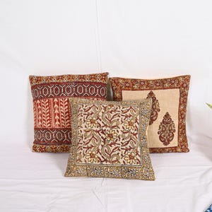 May include: Three square throw pillows with intricate floral and geometric patterns in shades of red, brown, and cream. The pillows are arranged on a white surface, showcasing their detailed designs and textures. The pillows add a touch of bohemian style.