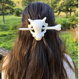 May include: A white hair clip featuring a skull design with a bone through the center. The hair accessory is worn in long, brown hair against a blurred green background.