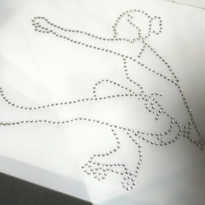 Op de afbeelding: Een witte stof met een zilveren strass-ontwerp van een kunstschaatser. Het ontwerp is omrand met strass.