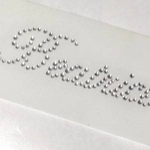 Puede incluir: Una superficie blanca con la palabra "París" escrita en pedrería plateada.