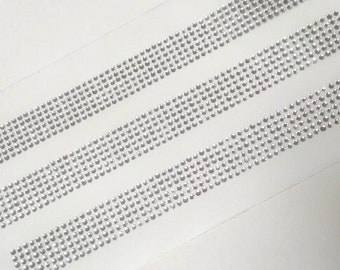 May include: Three rows of silver rhinestones on a white background. The rhinestones are arranged in a straight line and are evenly spaced.