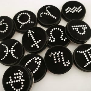 Op de afbeelding: Een verzameling ronde zwarte knopen, elk voorzien van een uniek sterrenbeeldsymbool gemaakt van kleine, heldere, reflecterende strass-steentjes. De set bevat diverse astrologische tekens die in een groep zijn gerangschikt.