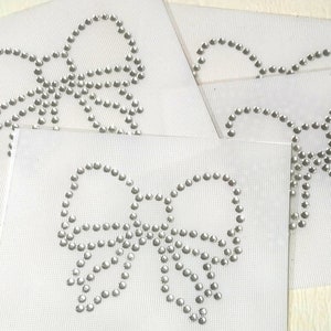 May include: Four white fabric squares with silver rhinestone bows. Each bow is made of small, round rhinestones.