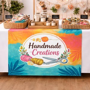 May include: A table display with a white tablecloth and a colorful runner featuring the text "Handmade Creations." Baskets hold various items, and small signs indicate product types. The runner has a watercolor design with spools of thread and scissors.
