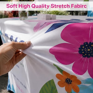 May include: A white stretch fabric table cover featuring a vibrant floral pattern with large pink and orange flowers and green leaves. The fabric displays the text Soft High Quality Stretch Fabirc at the top and includes additional printed text.