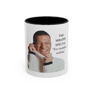 Puede incluir: Una taza de cerámica blanca con interior y borde negros, que presenta una impresión fotográfica de una persona sonriente con un suéter de punto blanco. El texto en la taza dice: THE MBAPPÉ SPECIAL "For example... nothing."