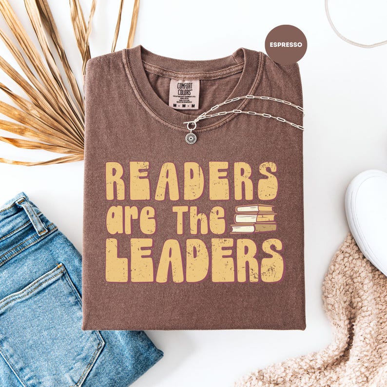 May include: A brown t-shirt with the text "READERS are The LEADERS" in a retro font. The shirt has a crew neck and short sleeves. A silver necklace with a pendant is draped across the shirt. A pair of blue jeans and a white shoe are visible.
