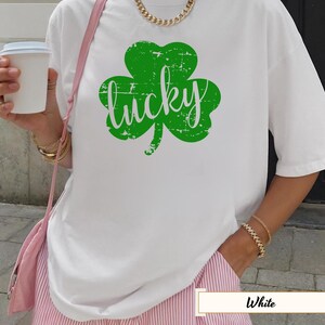 Könnte beinhalten: Weißes T-Shirt mit einem großen grünen Kleeblatt und dem Wort "lucky" in verblichener Schrift. Das Shirt wird mit einer rosa-weiß gestreiften Hose getragen. Eine goldene Halskette und Armbänder sind sichtbar.
