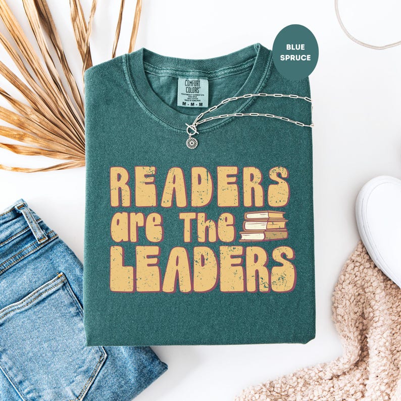 May include: A blue spruce t-shirt with the text "READERS are The LEADERS" in a retro font. The shirt has a crew neck and short sleeves. A silver necklace with a sun pendant rests on the shirt. The shirt is folded on a white surface.
