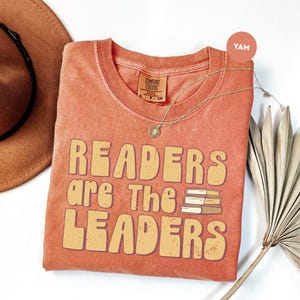 May include: Orange Comfort Colors t-shirt with the text "READERS are The LEADERS" in a retro font. The shirt features a stack of books graphic. A gold necklace and a brown hat are also visible.