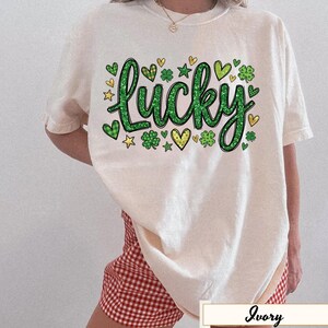 Könnte beinhalten: Cremefarbenes T-Shirt mit dem Wort "Lucky" in grünem Glitzer-Schriftzug. Das Design umfasst grüne Kleeblätter, Herzen und Sterne. Das Shirt wird mit rot-weiß karierten Shorts kombiniert.