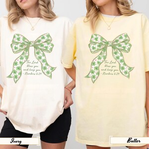 Könnte beinhalten: Zwei T-Shirts, eines in Elfenbein und eines in Buttergelb. Jedes Shirt hat ein großes grünes Schleifendesign mit Kleeblättern und dem Text "The Lord bless you and keep you - Numbers 6:24". Die Shirts haben eine lockere Passform und kurze Ärmel.