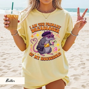 May include: Butter-colored t-shirt with the text "I AM THE PRIMARY BREADLOSER OF MY HOUSEHOLD." The graphic features a cartoon rat wearing sunglasses, a beret, and holding a cocktail. The rat has a purse and a gold bar. The shirt is a relaxed fit.