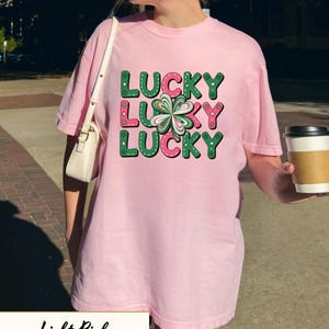 Op de afbeelding: Lichtroze T-shirt met het woord "LUCKY" in groene en roze blokletters, en een klaverblad-afbeelding. Het shirt heeft een lichtroze kleur.