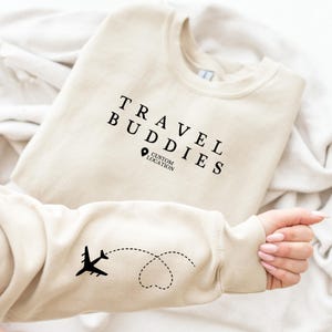 May include: A cream-coloured sweatshirt with the text "TRAVEL BUDDIES" and a location pin graphic printed in black. The sleeve displays a black airplane graphic with a dotted heart-shaped trail. The sweatshirt is folded on a white surface.