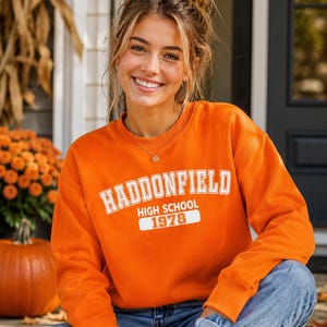 Könnte beinhalten: Ein leuchtend orangefarbenes Sweatshirt mit Rundhalsausschnitt und weißem Aufdruck HADDONFIELD HIGH SCHOOL 1978. Das Kleidungsstück hat lange Ärmel, einen gerippten Kragen und gerippte Bündchen und bietet eine klassische Passform für den Alltag.