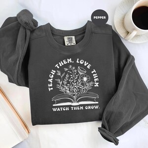 May include: A dark gray sweatshirt with the words "Teach Them, Love Them, Watch Them Grow" in a white, circular design. The design features an open book with floral illustrations. A cup of coffee sits in the background.