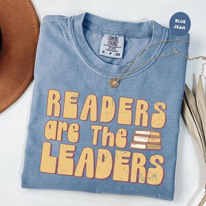 May include: Blue jean-colored t-shirt with the text "READERS are The LEADERS" in yellow letters. The shirt has a crew neck and short sleeves. A gold necklace with a pendant is draped across the shirt.