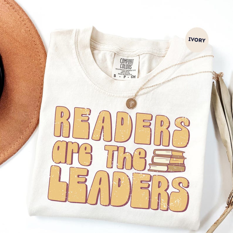 May include: Ivory-colored t-shirt with the text "READERS are The LEADERS" in a retro font. The design includes a stack of books. A gold necklace and a brown hat are also visible.