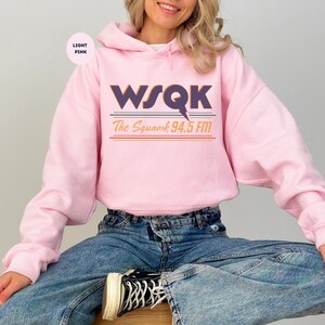 May include: Light pink hoodie with the text "WSQK The Squawk 94.5 FM" in purple, blue, and orange. The hoodie has a drawstring hood and long sleeves. The model is wearing blue jeans.