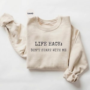 May include: Sand-colored sweatshirt with the text "LIFE HACK: DON'T START WITH ME" in black. The sweatshirt has a crew neck and long sleeves with rolled cuffs. The fabric appears soft and comfortable.