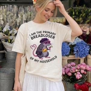May include: Cream-colored t-shirt with the text "I AM THE PRIMARY BREADLOSER OF MY HOUSEHOLD." The graphic features a cartoon rat wearing sunglasses, a beret, and holding a cocktail. The rat has a purse and a heart-shaped tail.