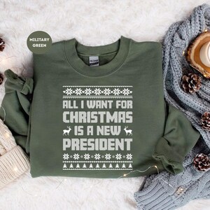 以下が含まれることがあります： 「ALL I WANT FOR CHRISTMAS IS A NEW PRESIDENT」の白文字がプリントされたミリタリーグリーンのスウェットシャツ。雪の結晶とトナカイのデザイン。クリーム色のニット帽とグレーのケーブル編みのスカーフも画像にあります。