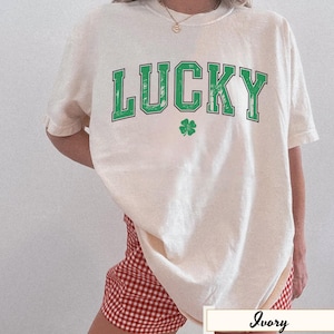 Könnte beinhalten: Elfenbeinfarbenes T-Shirt mit dem Wort "LUCKY" in grünen Blockbuchstaben und einem grünen Kleeblatt. Das Shirt hat eine lockere Passform und wird mit rot-weiß karierten Shorts kombiniert. Das Shirt ist ein lässiger Stil.