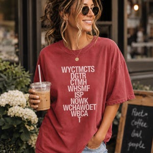 May include: A brick red short-sleeved crew neck t-shirt featuring white text printed in a vertical list format. The text reads: WYCTMCTS, DGTR, CTMH, WHSMF, ISP, NOWK, WCHAWOET, WBCA, T. The shirt has a relaxed fit and a slightly faded, worn-in appearance.
