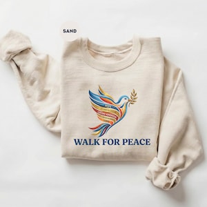 May include: A sand-colored sweatshirt featuring a colorful dove graphic holding an olive branch. The dove's wings are a rainbow of colors, with the words "HOPE, COMPASSION, LOVING KINDNESS, HARMONY, LOVE" on its body. The words "WALK FOR PEACE" are printed below.