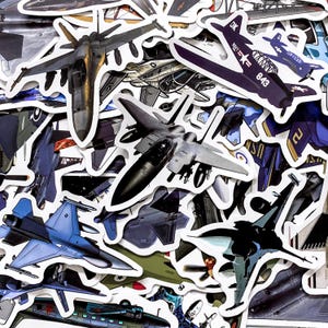 May include: A selection of airplane stickers in shades of blue, grey, and black. The stickers feature various fighter jets and military aircraft designs, each with a white border. Some stickers include text and numbers.