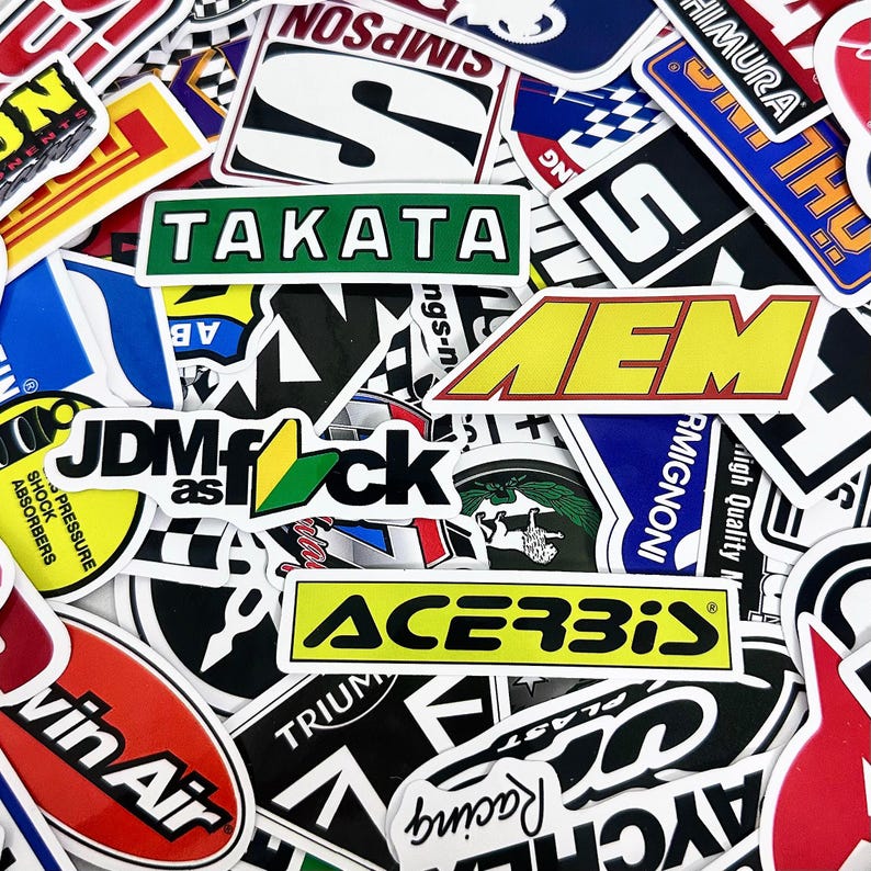May include: A collection of colorful stickers with various logos and text. The stickers feature brands like TAKATA, AEM, and ACERBIS, with a mix of shapes and designs. The stickers are arranged closely together.