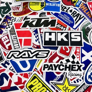 May include: A collection of colorful stickers with various logos and text. The stickers feature brands like HKS, RAYS, and PAYCHEX Racing. The stickers are in various shapes and sizes, with a mix of bold colors and fonts.